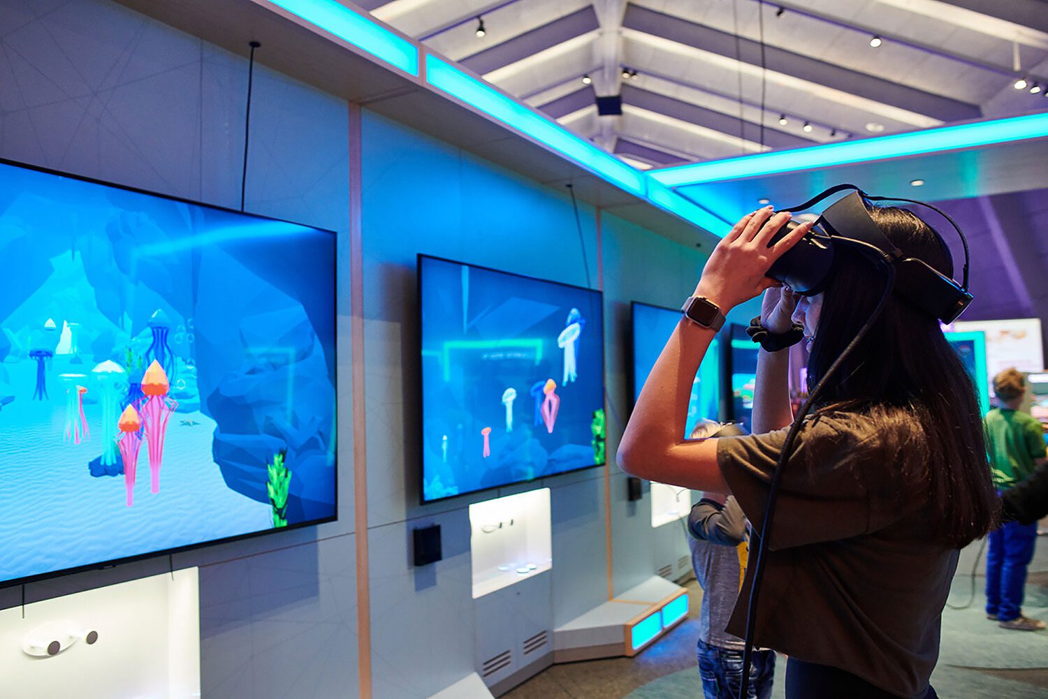 A girl with long brown hair puts on a virtual reality headset and examines a number of screens that show colorful jellyfish on them.